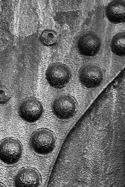 Black and white detail photograph of rusty metal surface with a curved pattern of big rivets.