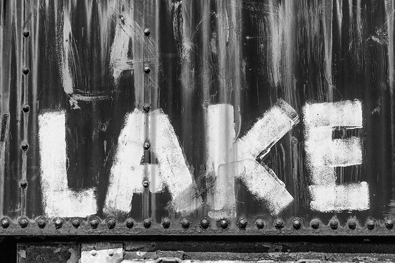 Black and white detail photograph of the word "LAKE" painted with a brush onto the side of a rusty, abandoned train car.