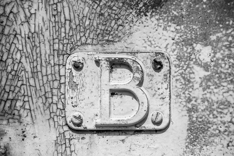 Black and white detail photograph of metal plate with a letter "B" bolted onto the cracked and peeling side of a rusty, abandoned railroad train car.