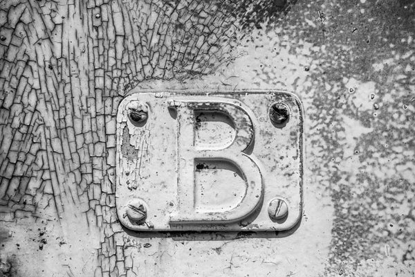 Black and white detail photograph of metal plate with a letter "B" bolted onto the cracked and peeling side of a rusty, abandoned railroad train car.
