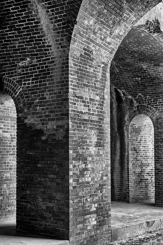 Black and white photograph of thick, brick columns and arches inside an old fort, but resembling something like a castle or dungeon.