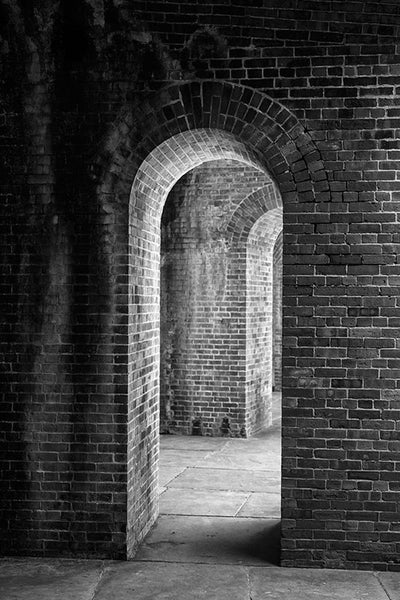 Black and white photograph of thick, brick walls with a series of repeated arched doorways, captured in dramatic lighting.