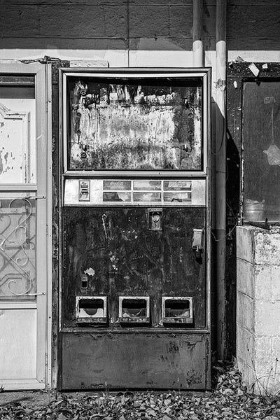 Black and white photograph of a textured, broken, and abandoned vending machine found along the side of a country road.