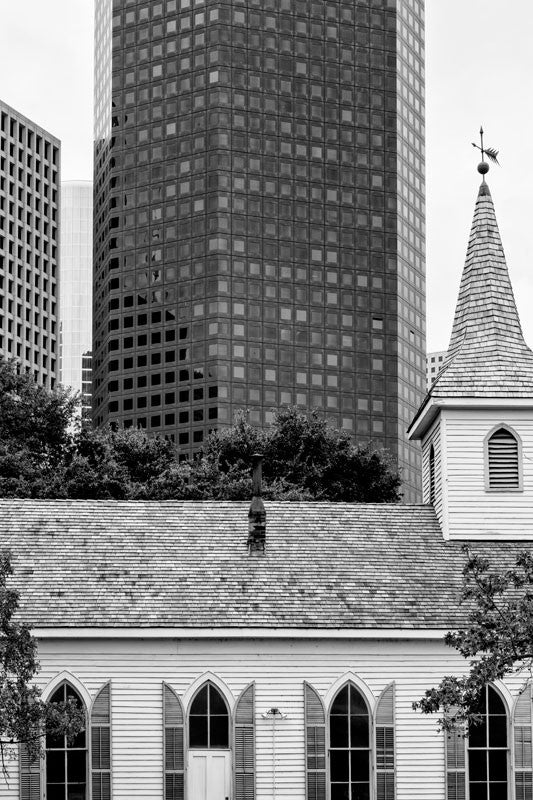 Black and white photograph of a small wooden historic church among the towering glass skyscrapers of downtown Houston.