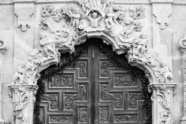 Black and white photograph of of the beautifully ornate hand-carved wooden doors at Mission San Jose in San Antonio, Texas. Around the carved doors is a framework of carved limestone reliefs featuring cherubic angels.