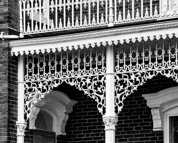 Black and white photograph of ornate, white lacy cast iron on the porch of a big Victorian house in the south. The ironwork is so intricate, it resembles like a white veil.
