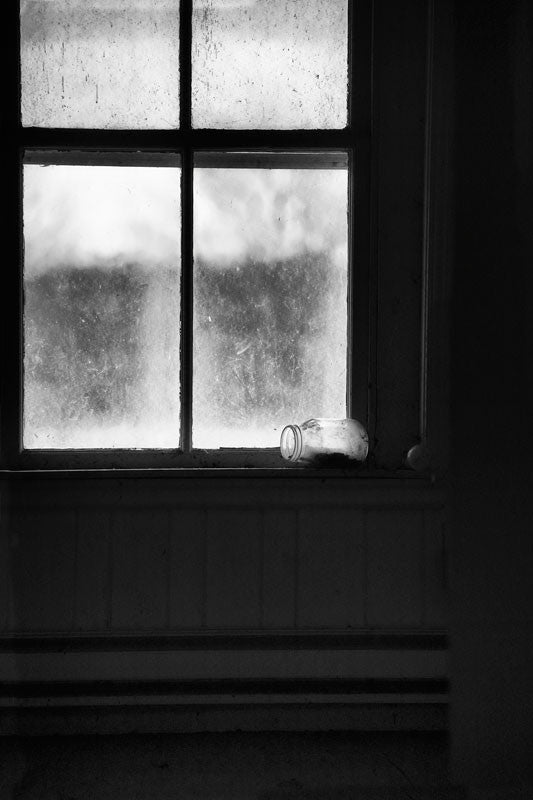 Black and white photograph of the dark interior of an abandoned old house, with frosty windows, and an overturned glass jar on the window sill, photographed as it was found.