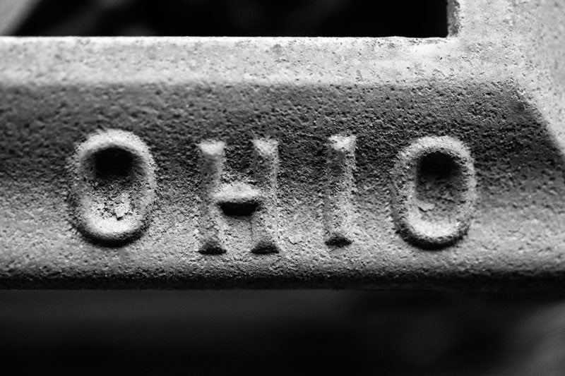 Black and white photograph of the word "Ohio" stamped into the metal of a rusty antique piece of farm equipment.