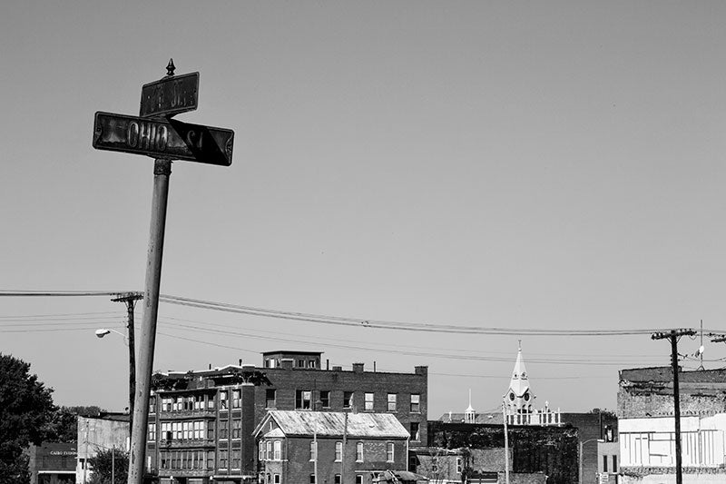 Black and white photograph of mostly abandoned Cairo, Illinois, with a rusty street sign in the foreground. Most of the buildings visible in this photograph are unoccupied and left to slowly decay.