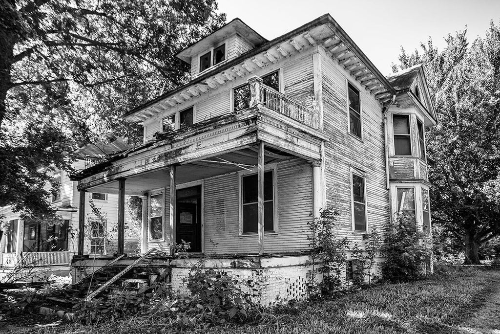 Black and white photograph of large, white house with peeling paint and broken woodwork, that has been abandoned and left standing wide open in a neighborhood.