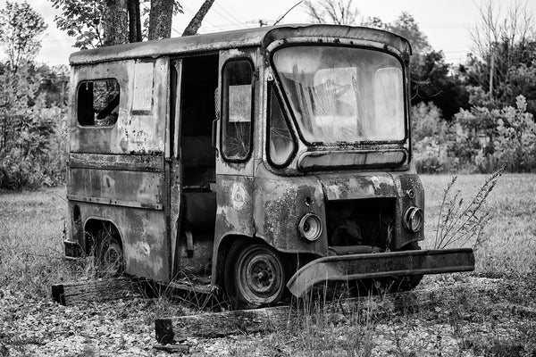 Black and white landscape photograph focused on a small rusty and abandoned Postal Service truck parked in a field. On its side is an old triangular Civil Defense logo, which says in a circle around the CD icon, "This is a unit of your Civil Defense."