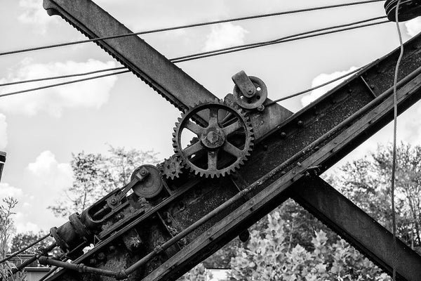 Black and white photograph of the gears and cables of a rusty, old steam shovel abandoned in a grassy rural pasture.
