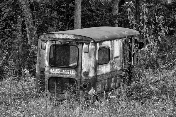 Black and white photograph of a rusty, old US Mail truck abandoned in a grassy field.