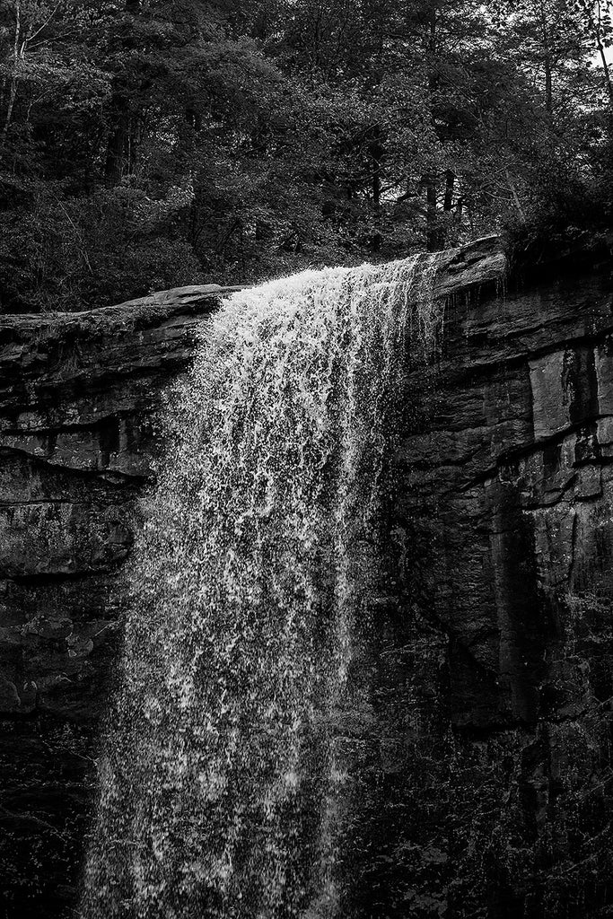 Black and white landscape photograph of a waterfall in dramatic low light, emphasizing the whitewater of the river spilling over the edge of the cliff. This photograph captures many of the individual water droplets in mid-air.