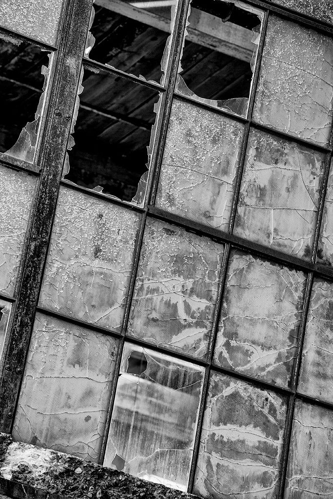 Black and white architectural detail photograph of a bank of windows in an abandoned factory, with some panes missing, and others beautifully textured, cracked, and peeling.