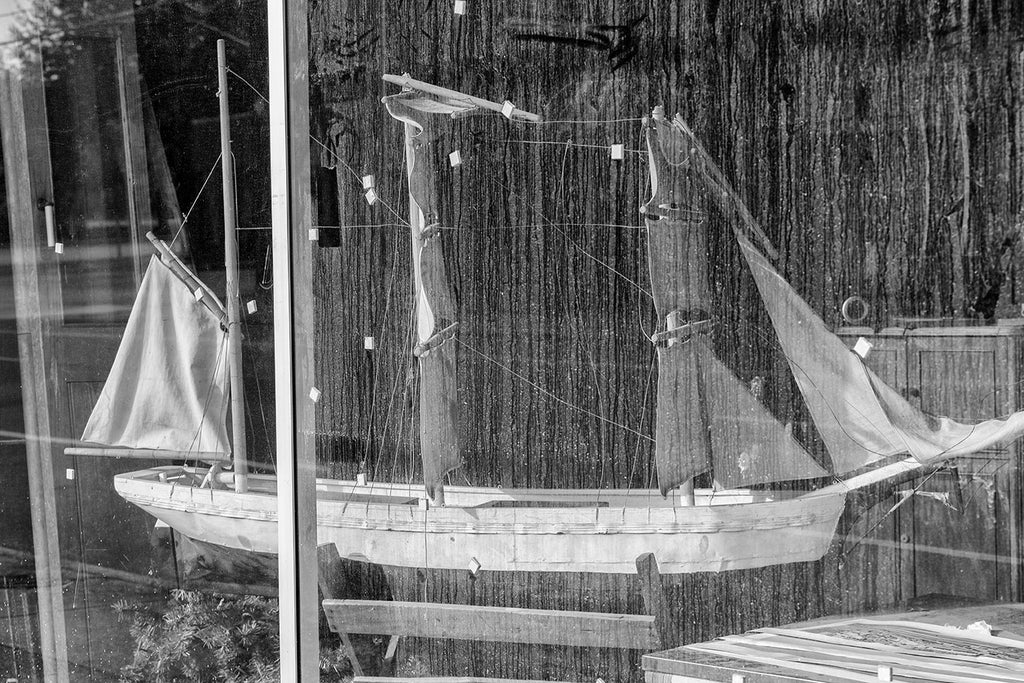 Black and white photograph of a model of a tall sailing ship displayed behind a dirty, streaked store window.