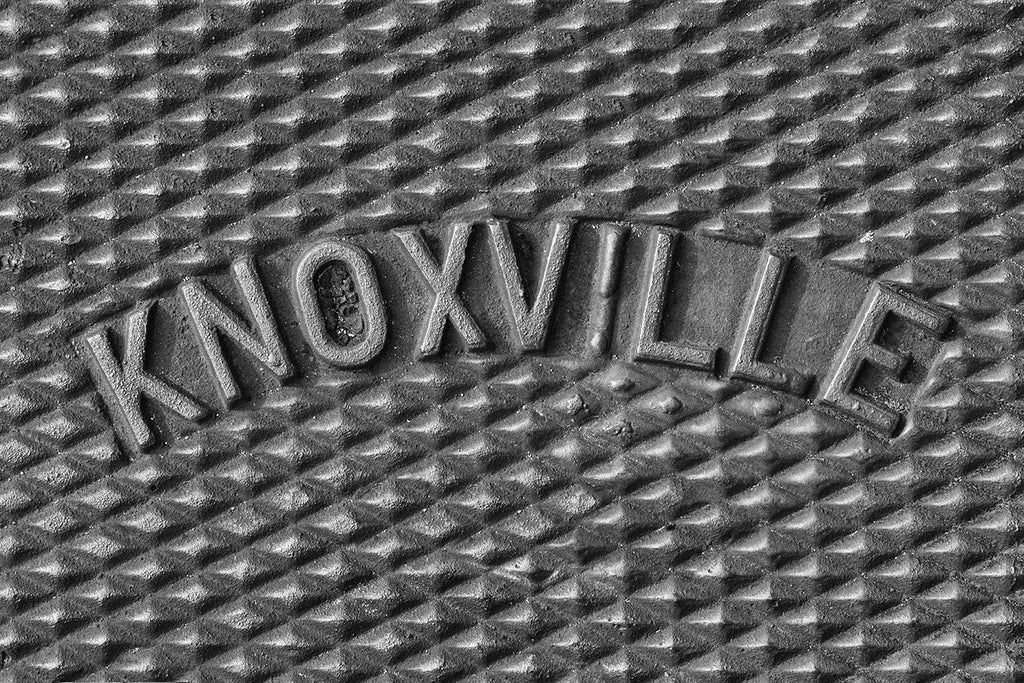 Black and white photograph of the word "Knoxville" curved across an iron utility cover on a sidewalk in Knoxville, Tennessee.