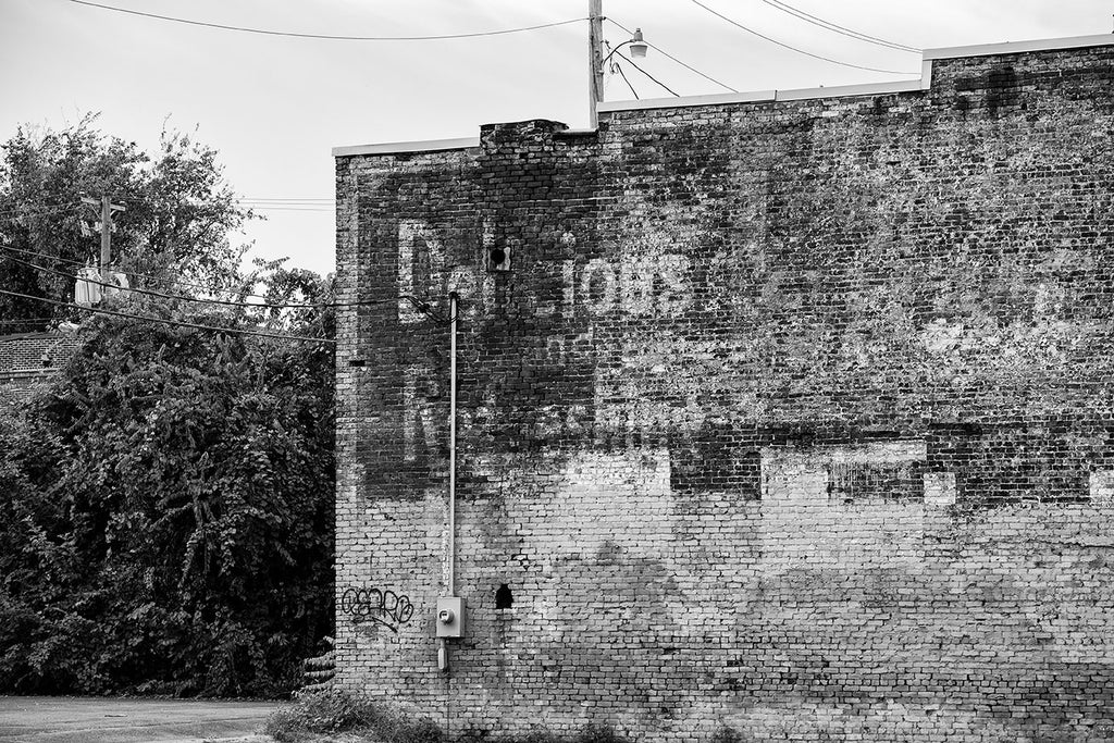 Black and white photograph of a fading, peeling ghost sign ad on the side of an old brick building. The most prominent legible word is "Delicious."