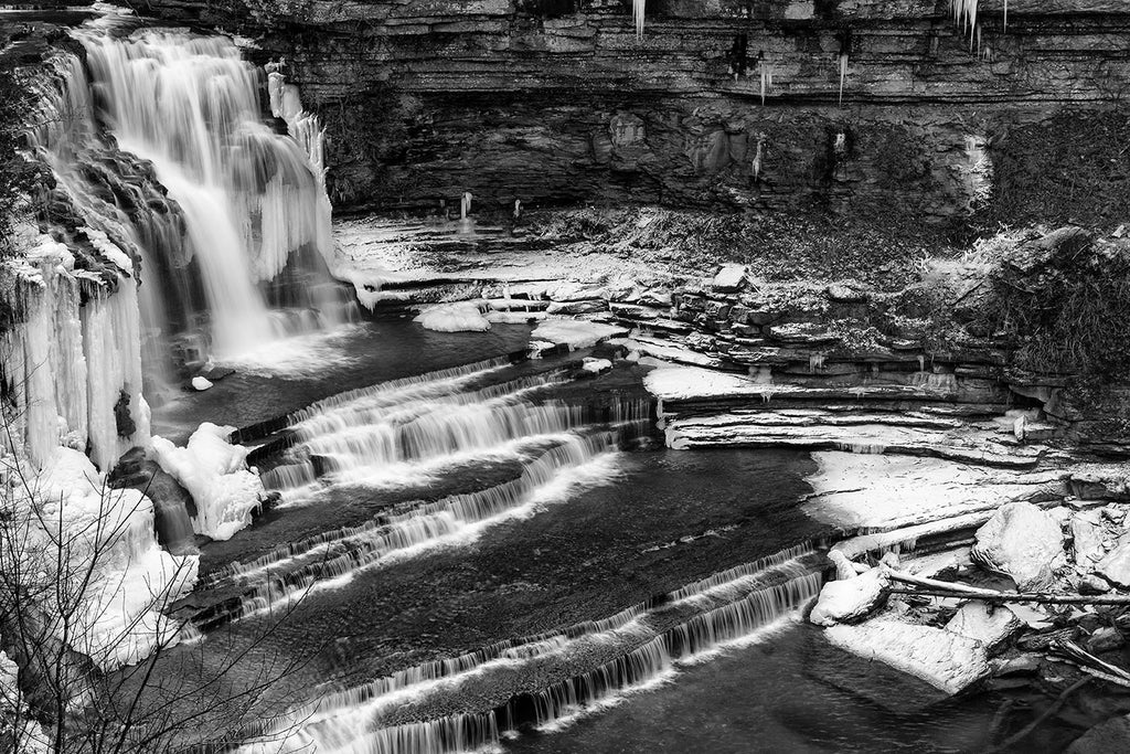 Black and white landscape photograph of icy winter waterfalls surrounded by snow and icicles seen from above.