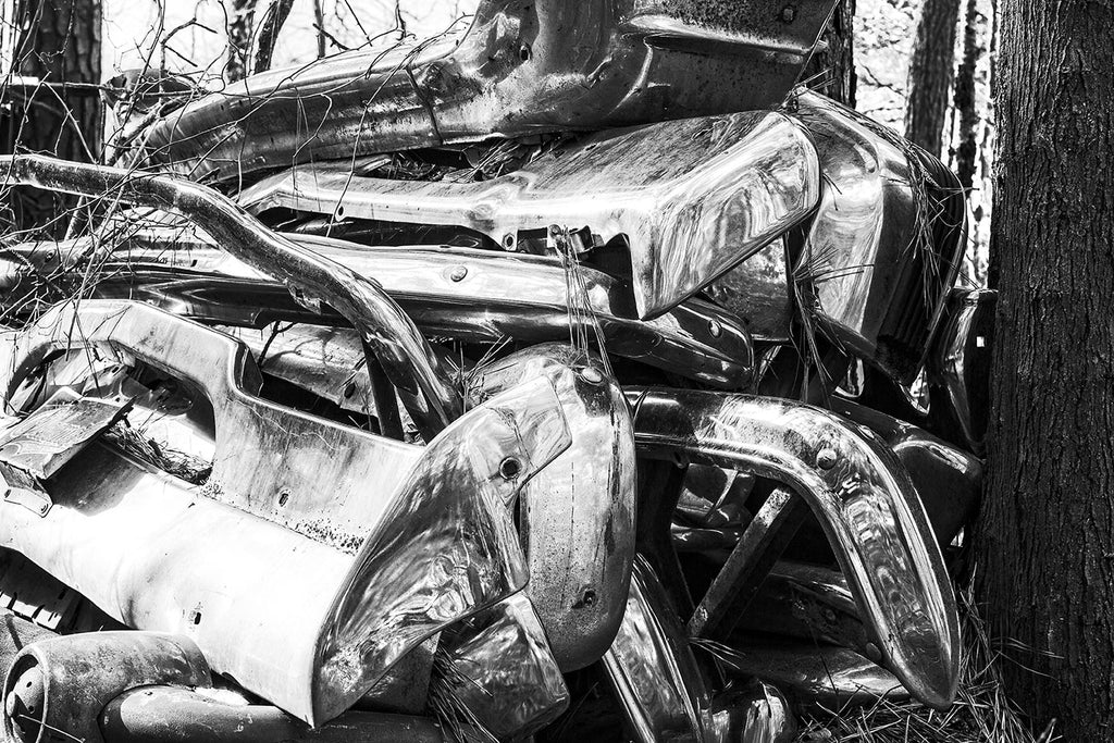 Black and white fine art photograph of big chrome bumpers from vintage American automobiles piled on top of each other in the woods.