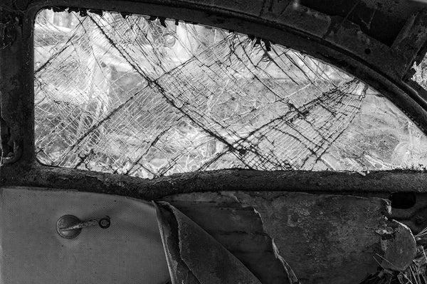 Black and white fine art photograph of the interior of an antique roadster, which has seen better days. The window is fractured in beautiful patterns, but the torn interior provides period context.
