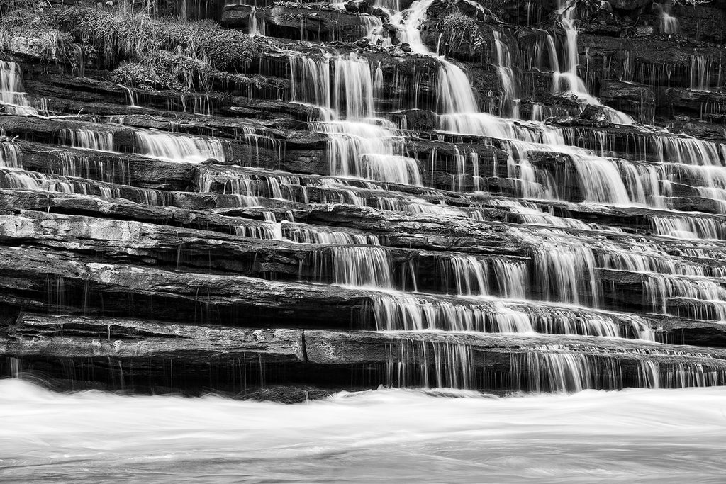 Black and white landscape photograph of a waterfall cascading down terraced rock ledges into a rushing river.