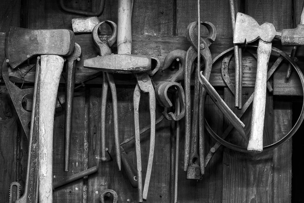 Black and white fine art photograph of a collection of rusty tools hanging on the wall of a working blacksmith shop.