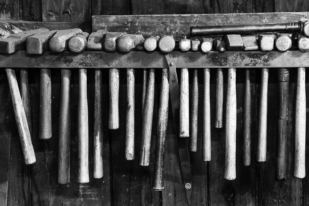 Black and white fine art photograph of a row of rusty hammers hanging on the wall of a working blacksmith shop.