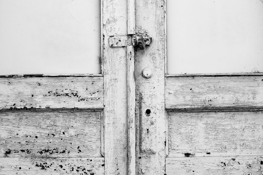 Black and white photograph of a cracks and chips showing dark through the aging white paint on the wooden doors of an abandoned building in a small town.