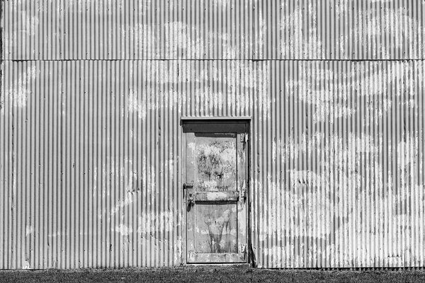 Black and white photograph of a tin industrial building with a grimy door set into a wall with strong vertical ridges.