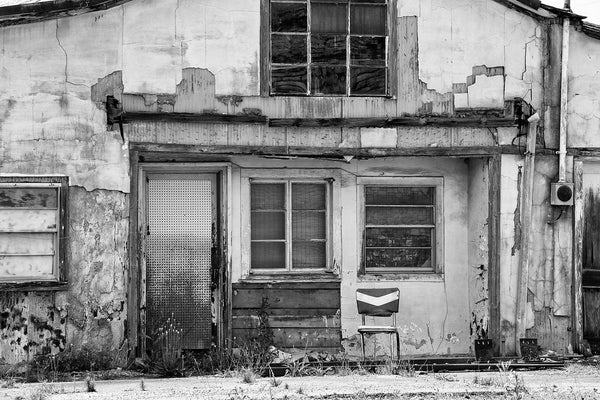 Black and white photograph of a shabby house with a vintage chair on the front porch.