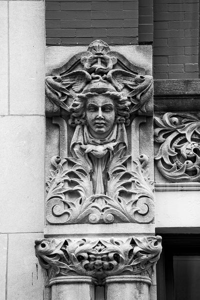 Black and white photograph of a carved face on the Drhumor Building (pronounced "drummer") on 48 Patton Avenue in downtown Asheville, North Carolina. Built in 1895, the building features a set of carved faces, believed to have been modeled by local citizens, created by English sculptor Frederick Miles, who also worked on the nearby Biltmore Estate.  This photograph is one of a set of six, designed to be displayed together.