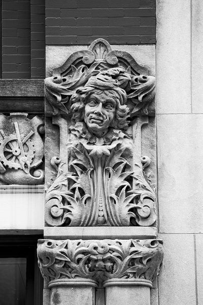 Black and white photograph of a man's face carved on the Drhumor Building (pronounced "drummer") on 48 Patton Avenue in downtown Asheville, North Carolina. Built in 1895, the building features a set of carved faces, believed to have been modeled by local citizens, created by English sculptor Frederick Miles, who also worked on the nearby Biltmore Estate. This photograph is one of a set of six, designed to be displayed together.