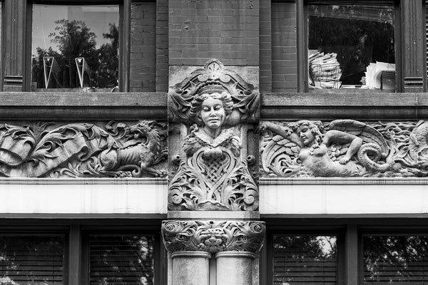 Black and white photograph of a fantastical carved frieze on the exterior of 48 Patton Avenue in downtown Asheville, North Carolina. The sweeping fantasy of the mythical creatures and iconic figures contrasts (and clashes) with the reality of the stacks of paperwork seen in the windows of the modern law office that inhabits the building.