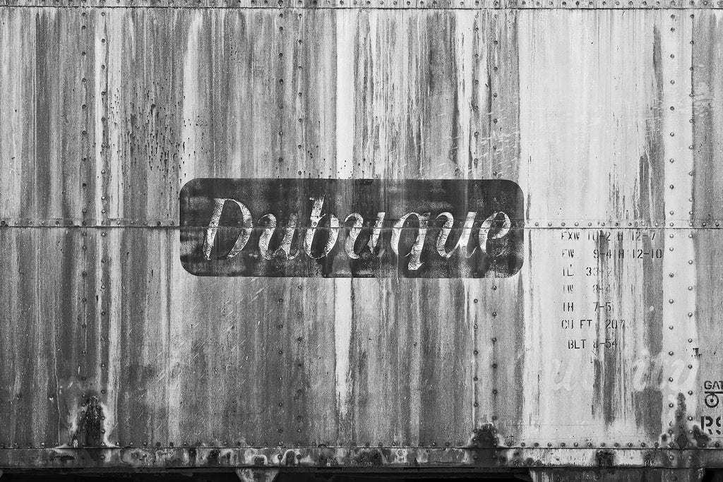 Black and white photograph of an abandoned antique railroad car with the word "Dubuque" painted on its side.