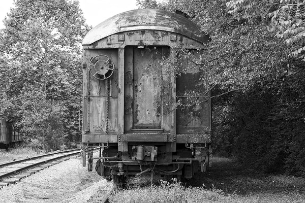 Black and white photograph of an abandoned and rusty old train car sitting on railroad tracks among the trees.