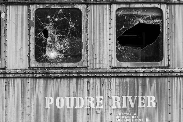 Black and white photograph of an abandoned railroad dining train car with shattered window glass. The side of the car displays the words "Poudre River," which runs through the US state of Colorado.