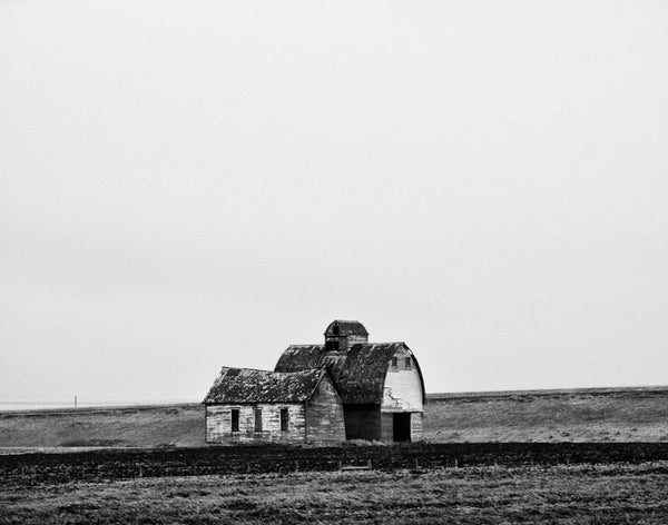 Black and white landscape photograph of an abandoned barn and farmhouse on the cold Minnesota prairie.