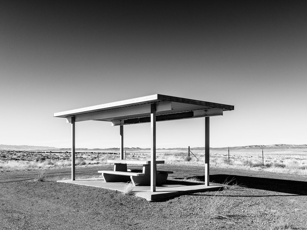 Black and white photograph of a roadside picnic table set amidst the vast and empty desert landscape of New Mexico.