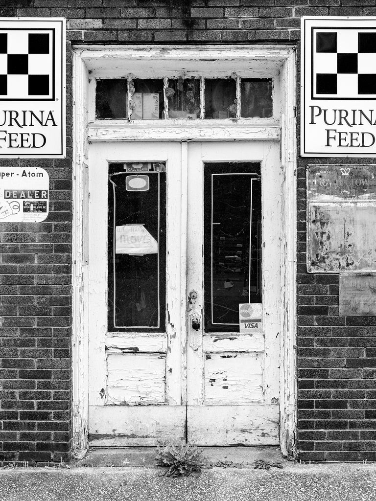 Black and white photograph of the front doors of a vacant old country feed store covered with the faded and rusting signs of agricultural product companies.