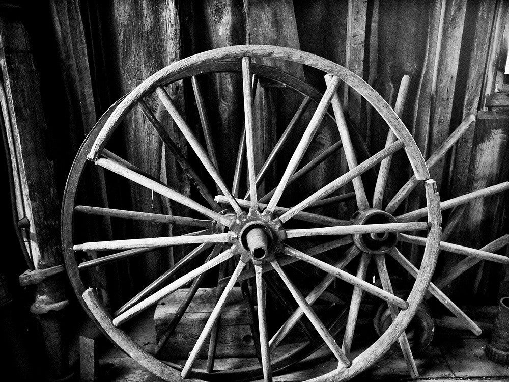 Black and white photograph of a broken antique wagon wheel found inside a wooden barn.