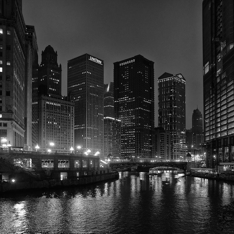 Black and white photograph of the downtown Chicago skyline at night, with city lights reflecting in the black Chicago River.