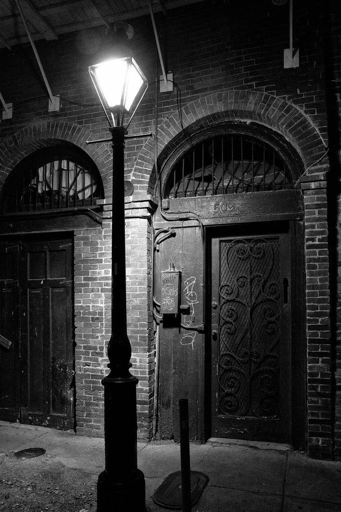 Black and white photograph of a New Orleans French Quarter street after dark illuminated by the light of a lamp post.