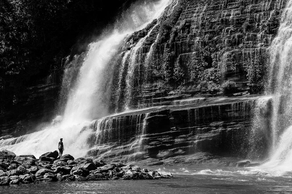 Moody black and white landscape photograph of dramatic waterfalls with a person in foreground providing a sense of scale.