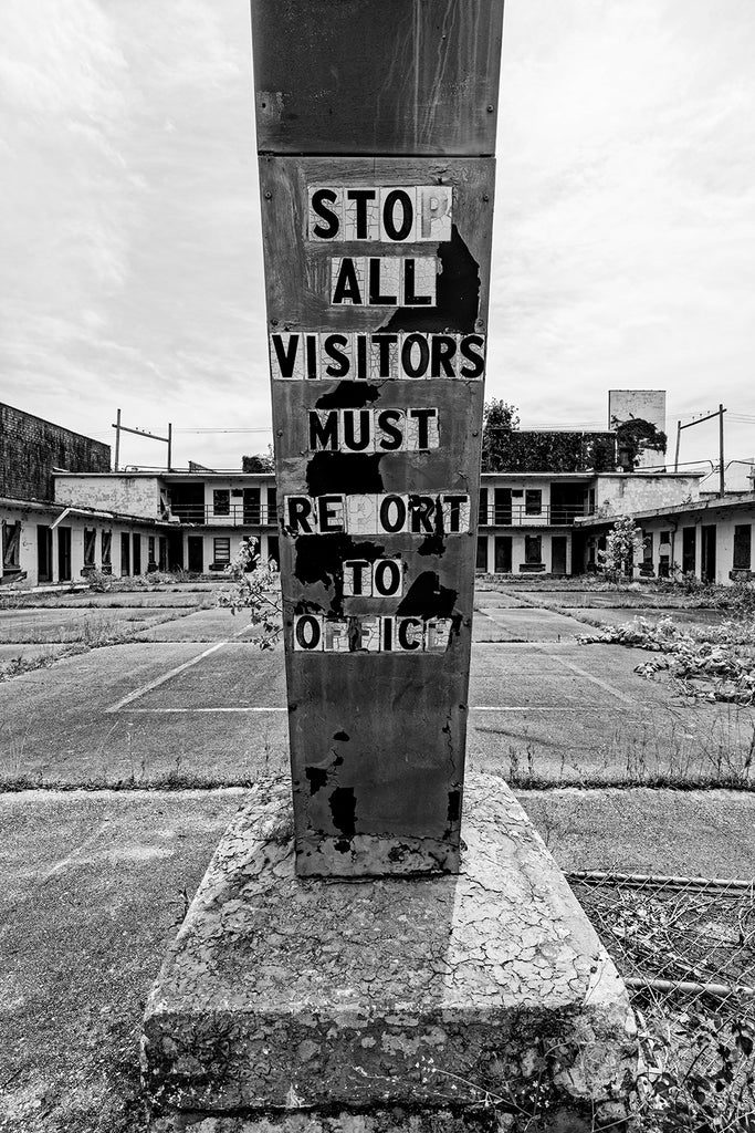 Black and white photograph of an abandoned motel with message on the post of its old sign that says "Stop All Visitors Must Report to Office."