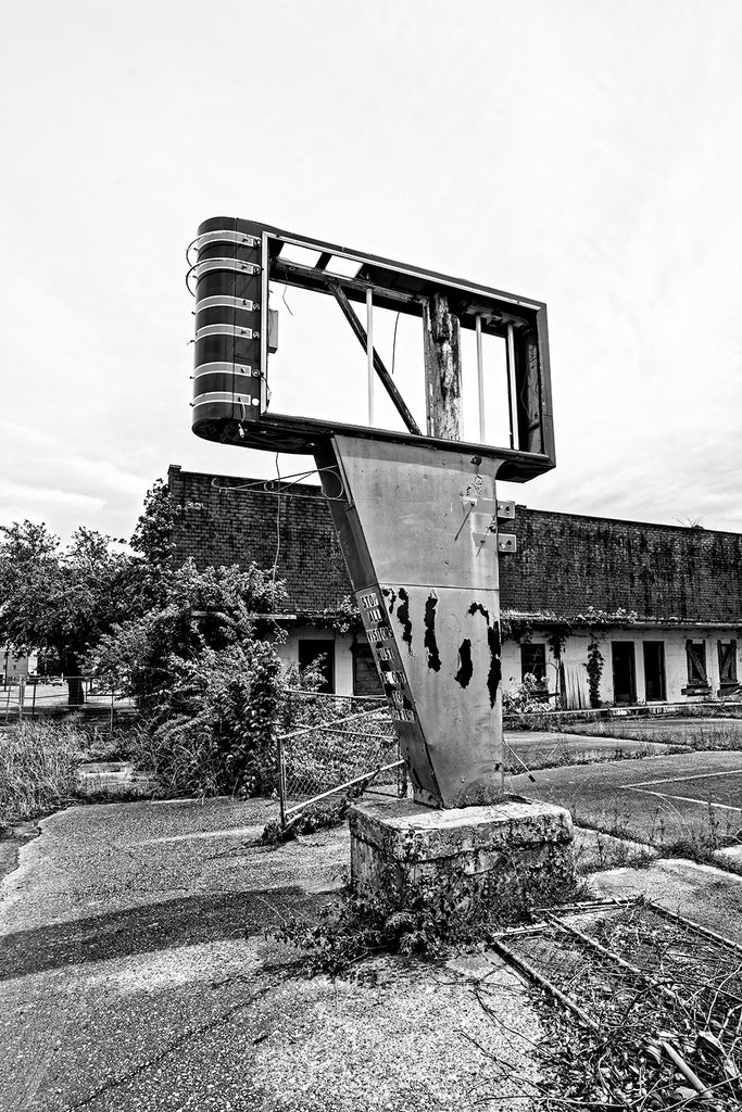 Black and white photograph of a broken old sign on the edge of a parking lot at an abandoned motel in the Mississippi Delta.