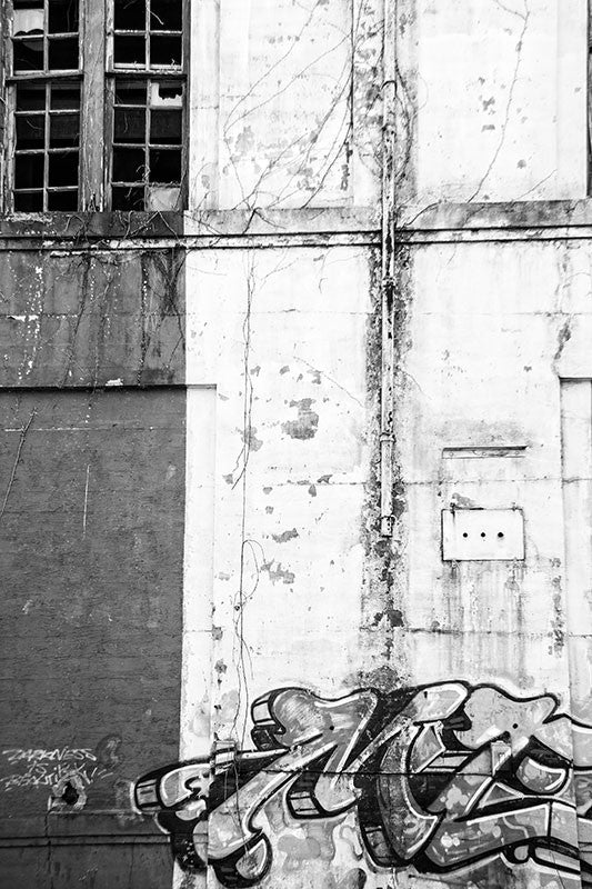 Black and white photograph of the wall of an abandoned building with graffiti and broken windows. Vertical composition.