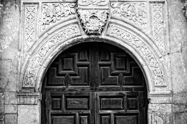 Black and white photograph of the arch over the entrance to the world-famous Alamo, cradle of Texas independence from Mexico in San Antonio, Texas. This architectural detail photograph illustrates the ornate stone carvings and textures of the building, which began existence as a Spanish mission in 1744, known originally as Misión San Antonio de Valero.