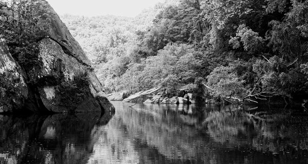 Black and white landscape photograph looking along a glassy river surrounded by giant rocks and forested hills.
