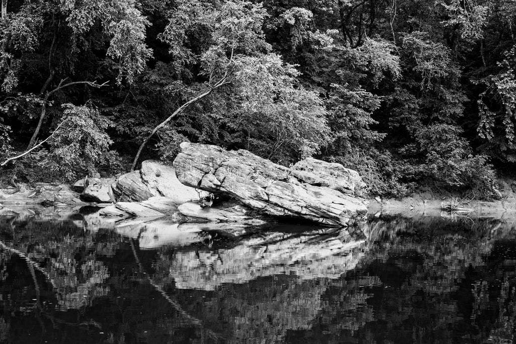 Black and white landscape photograph of large boulders reflecting into the placid surface of a slow-moving river. 
